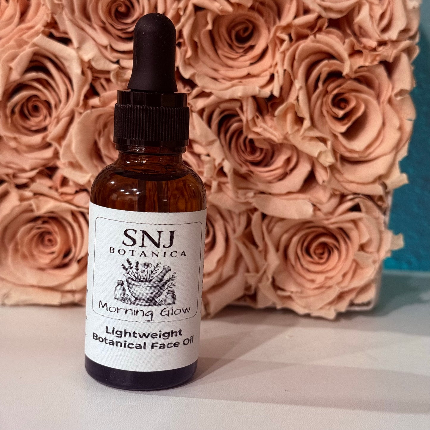 A bottle of SNJ Botanica Morning Glow Lightweight Botanical Face Oil with a black dropper cap, set against a background of dried peach-pink roses.
