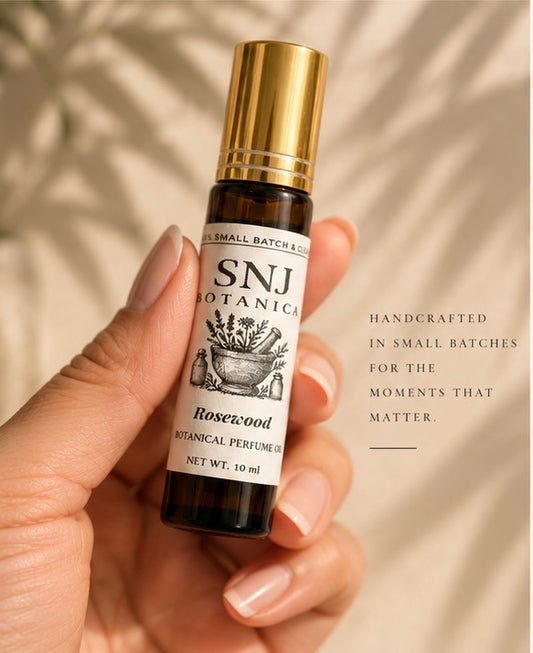 A 10 ml amber glass roll-on bottle of SNJ Botanicals Rosewood Botanical Perfume Oil with a shiny gold cap, styled being held by a hand.