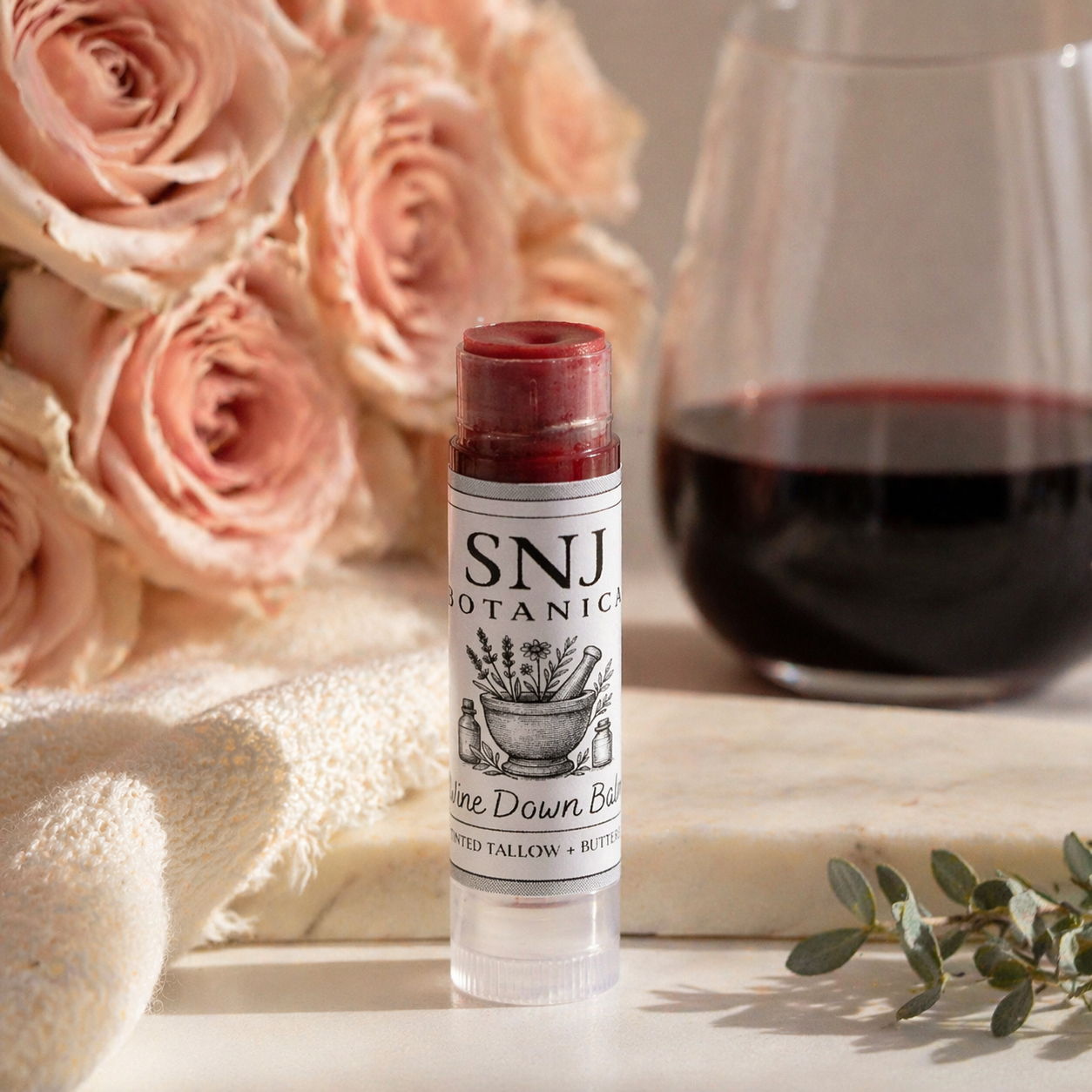 Lip balm with 'SNJ Botanics' branding in front of pink roses and a glass of red wine.