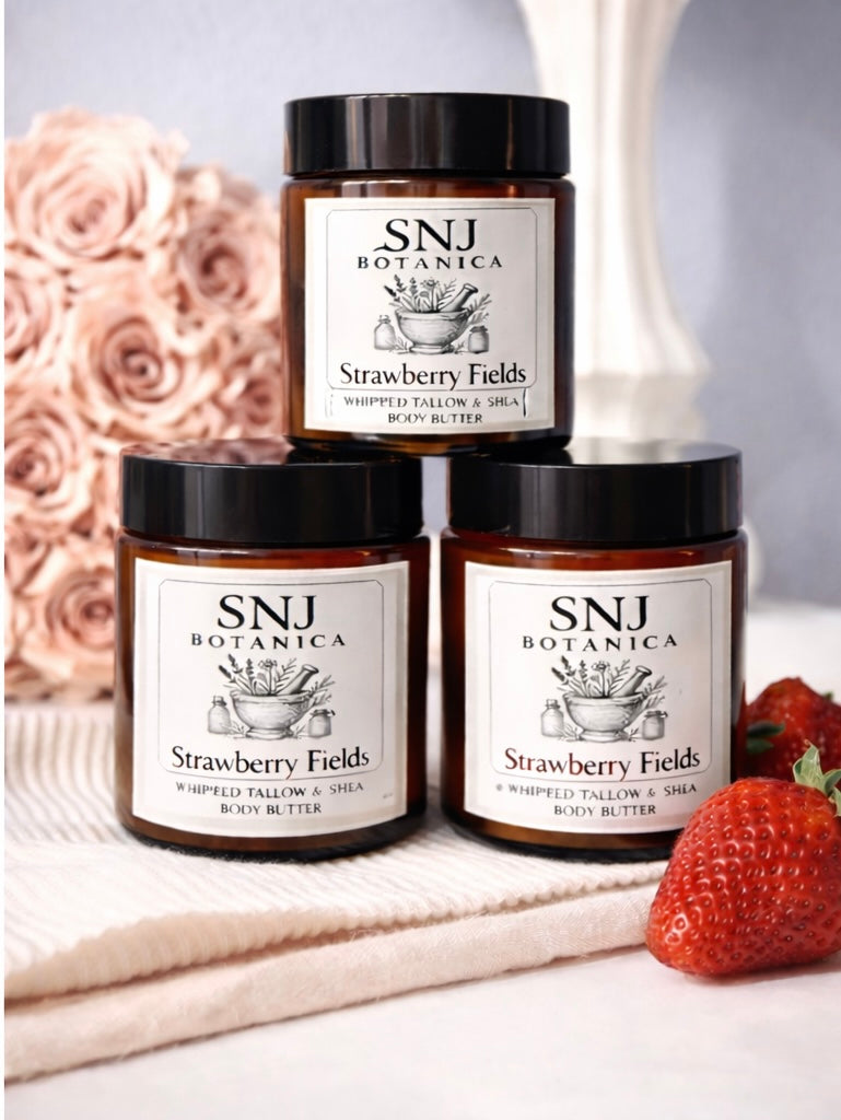 Three jars of SNJ Botanica Strawberry Fields body butter on a white surface with a strawberry and pink flowers in the background.