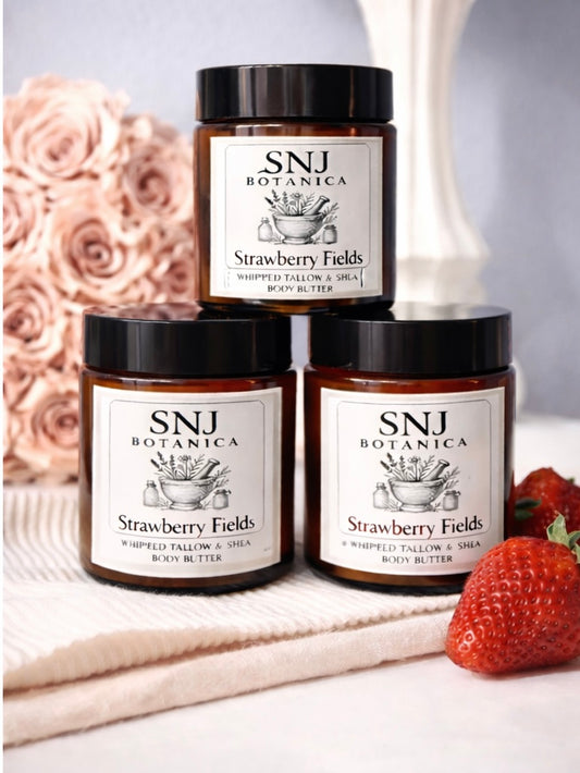 Three jars of SNJ Botanica Strawberry Fields body butter on a white surface with a strawberry and pink flowers in the background.
