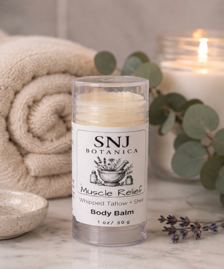 Body balm stick with 'SNJ Botanica' label on a marble surface with a candle and towel in the background.
