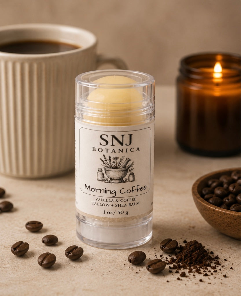 Balm stick with 'SNJ Botanica' label on a surface with coffee beans, a cup of coffee, and a candle.