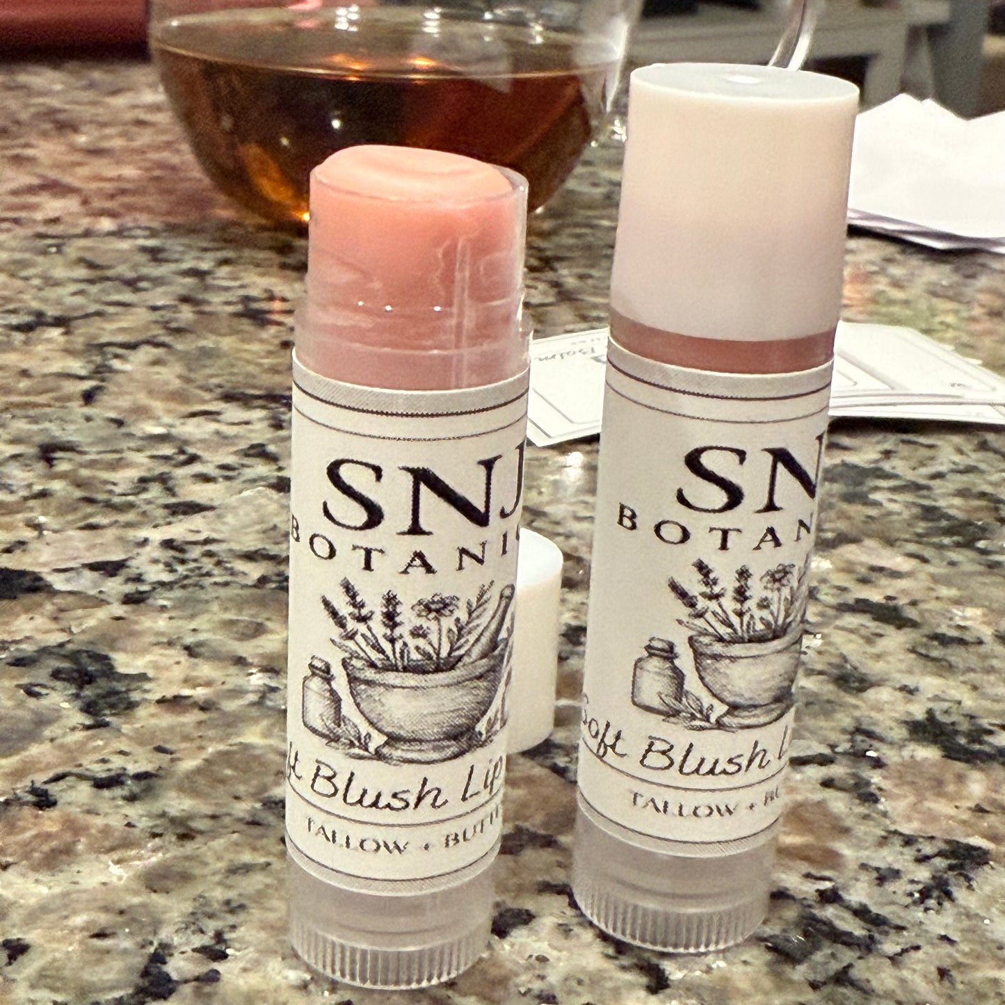 Two SN Botanical blush lip balms on a marble countertop with a glass of wine in the background.