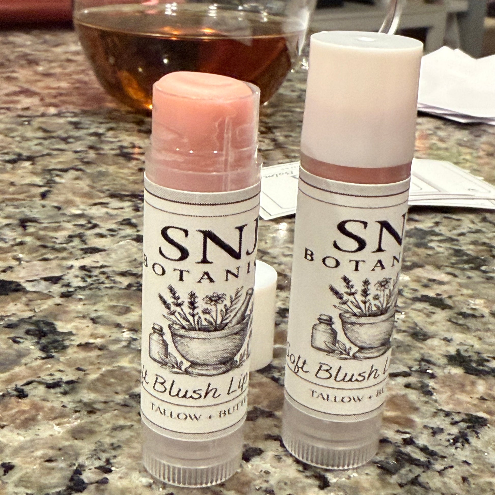 Two SN Botanical blush lip balms on a marble countertop with a glass of wine in the background.