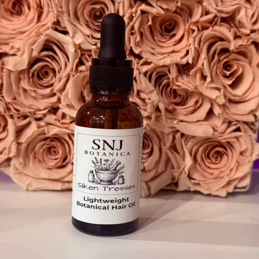 A bottle of SNJ Botanica Siken Tresses Lightweight Botanical Hair Oil with a black dropper cap, set against a background of dried beige roses.