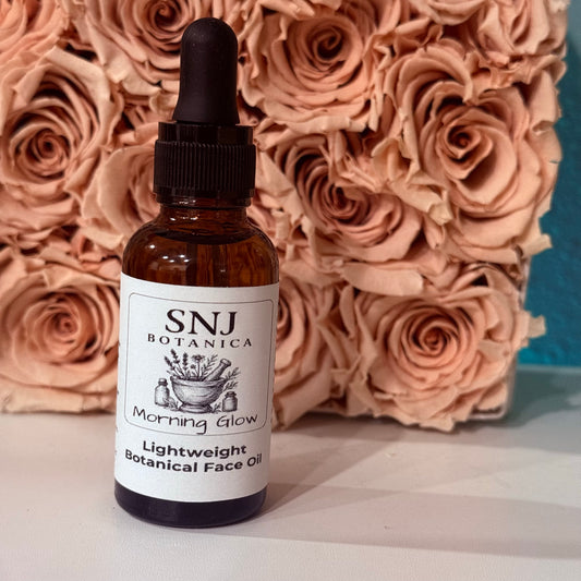 A bottle of SNJ Botanica Morning Glow Lightweight Botanical Face Oil with a black dropper cap, set against a background of dried peach-pink roses.