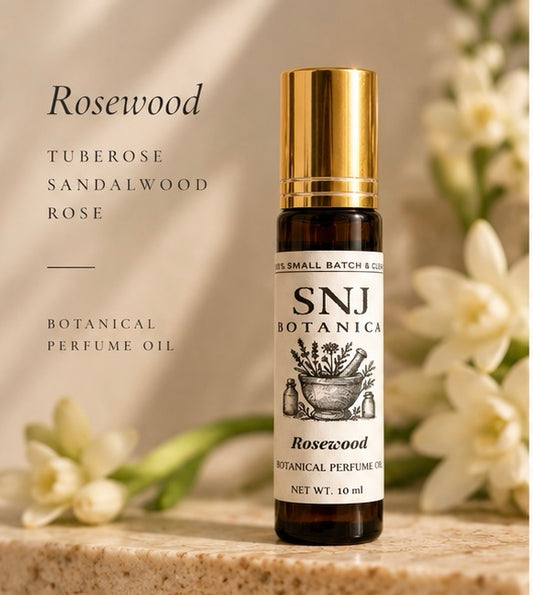 A 10 ml bottle of SNJ Botanicals Rosewood Botanical Perfume Oil with a gold cap, displayed on a stone surface with white flowers in the background and text on the left.
