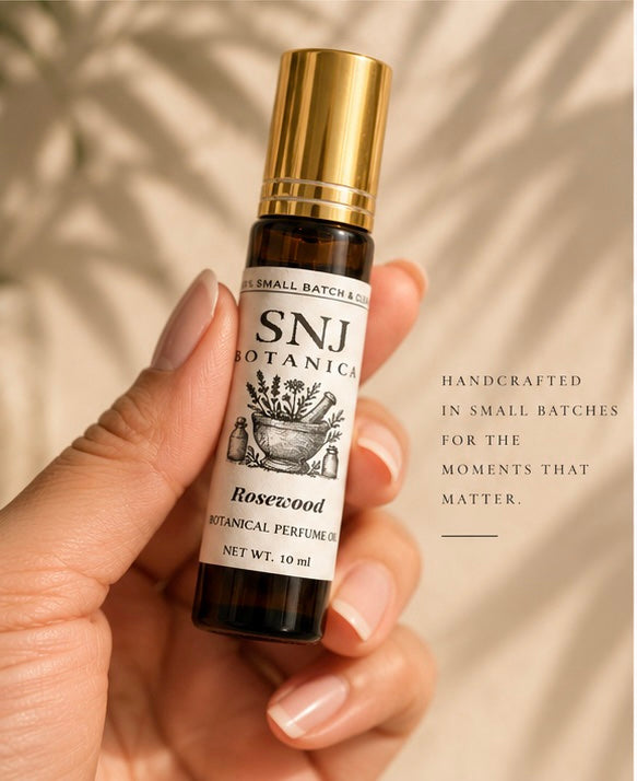 A 10 ml amber glass roll-on bottle of SNJ Botanicals Rosewood Botanical Perfume Oil with a shiny gold cap, styled being held by a hand. 