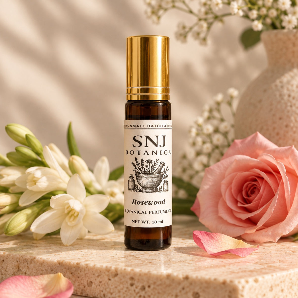 A 10 ml amber glass roll-on bottle of SNJ Botanicals Rosewood Botanical Perfume Oil with a shiny gold cap, styled on a stone surface with a pink rose, white flowers, and a textured vase in the background.